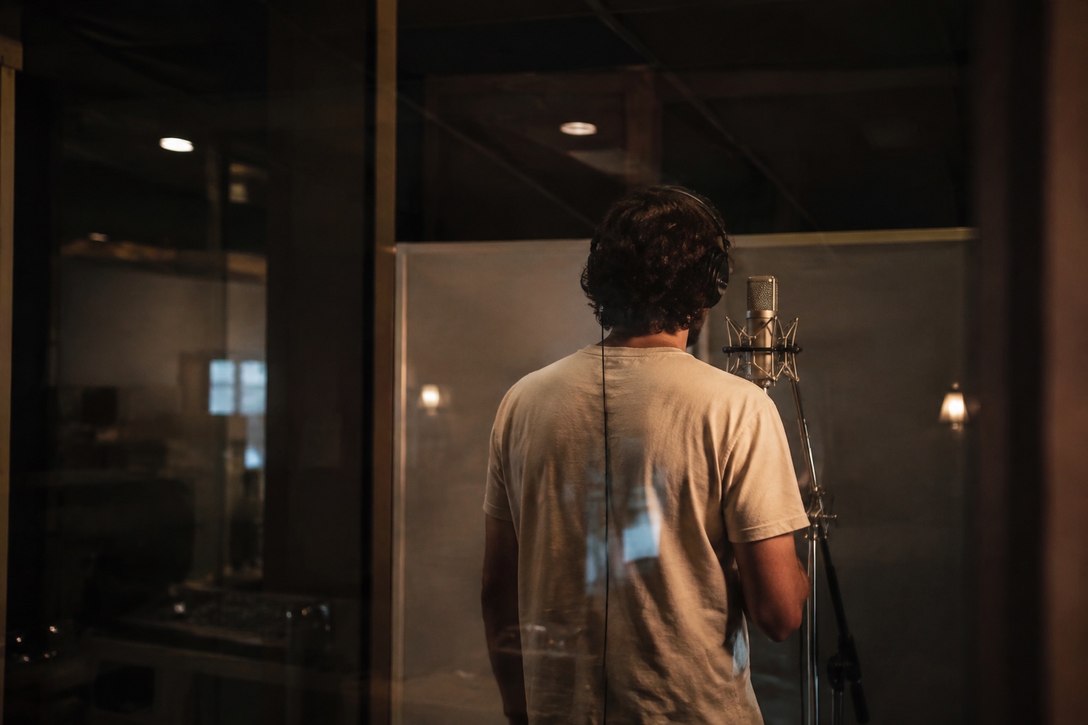 AI-generated fictional performer seen from behind through vocal-booth glass. Not Daniel, Kai or a documented AER session.